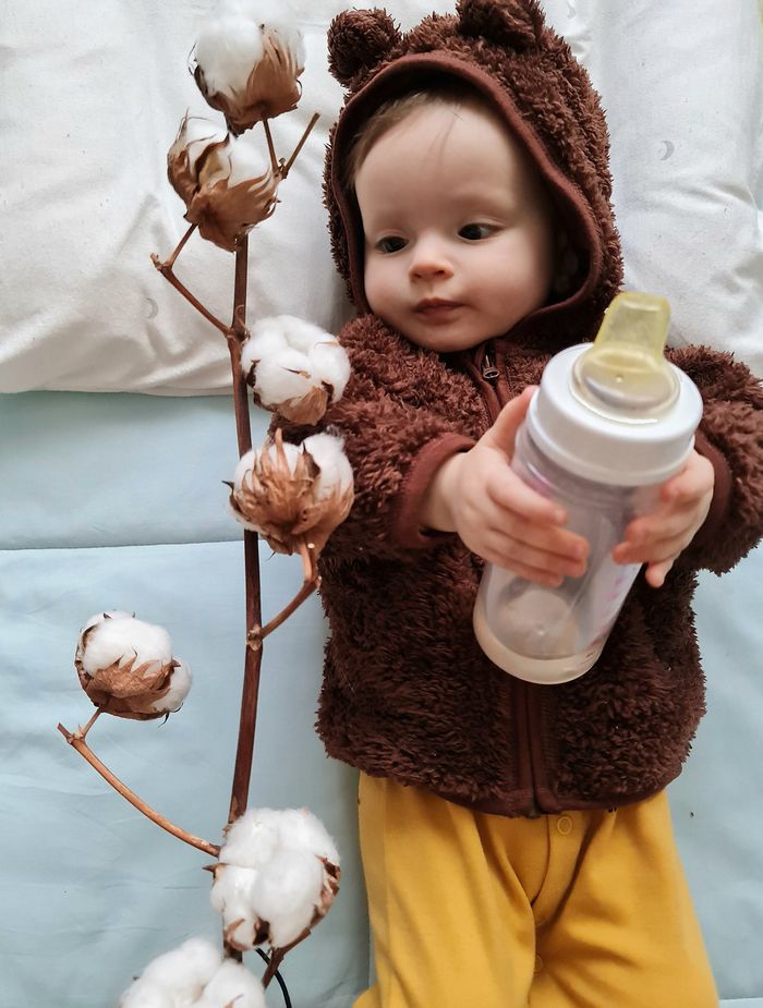 A cute baby in a teddy bear hoodie holds a bottle next to fluffy cotton branches.
