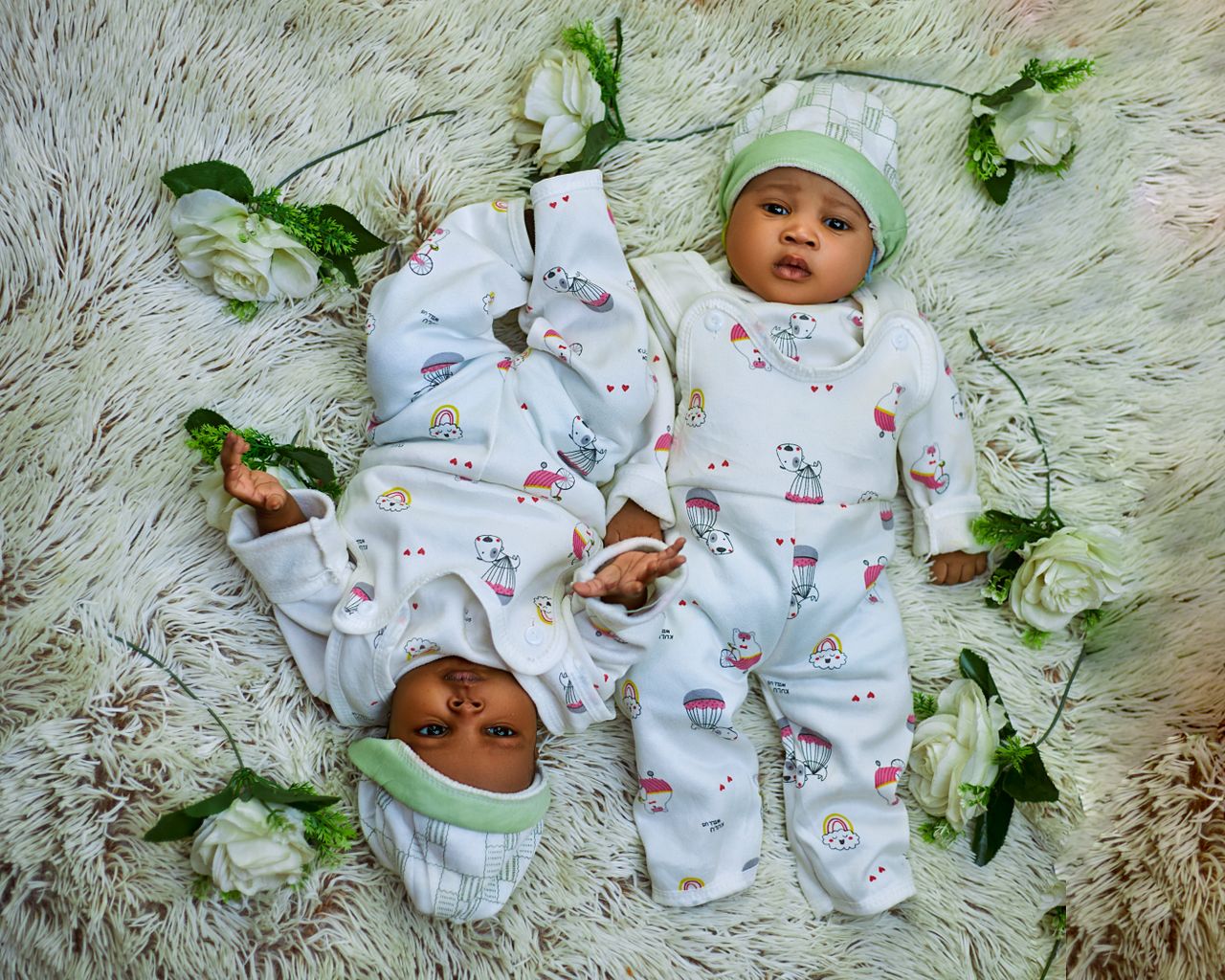 Cute twin babies in matching pajamas lying on a soft rug surrounded by white flowers.