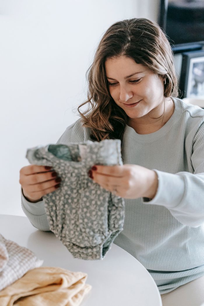 Positive female in casual wear folding baby clothes while sitting at white table with various garments in light room at home